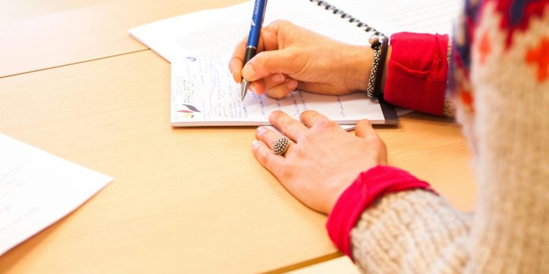 A woman writing notes in a notebook during a classroom lecture setting.