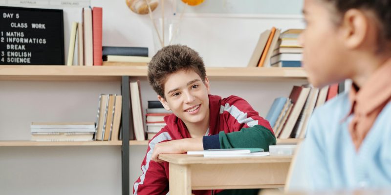 Teenagers smiling and interacting in a school classroom setting with books and educational materials.